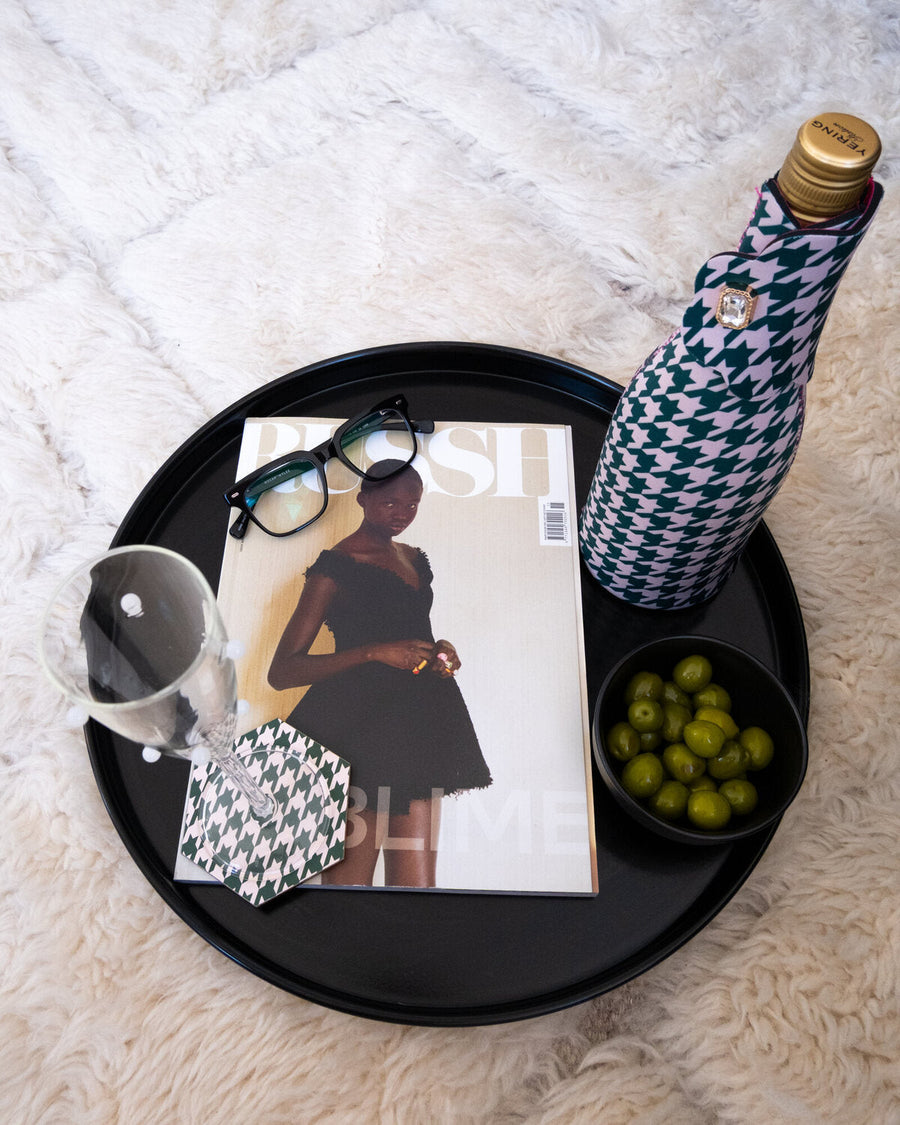 Black tray with a magazine, wine glass on a Chic Sips houndstooth coaster, wine bottle in a Chic Sips houndstooth bottle jacket, and a bowl of green olives on a plush white surface