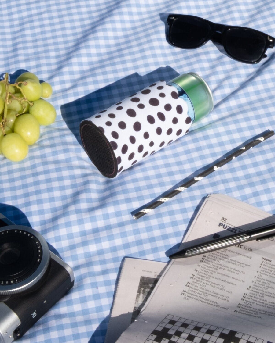 A styled flat-lay on a blue and white checkered blanket featuring a Chic Sips white can cooler with black polka dots, green grapes, black sunglasses, a silver and blacked camera, a striped Chic Sips straw, and a newspaper crossword.