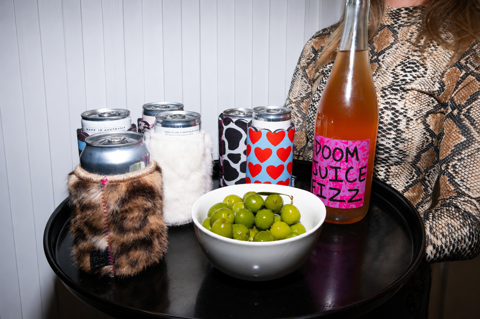 Assorted Chic Sips can sleeves displayed on a drinks tray with canned beverages, olives and a bottle of sparkling drink