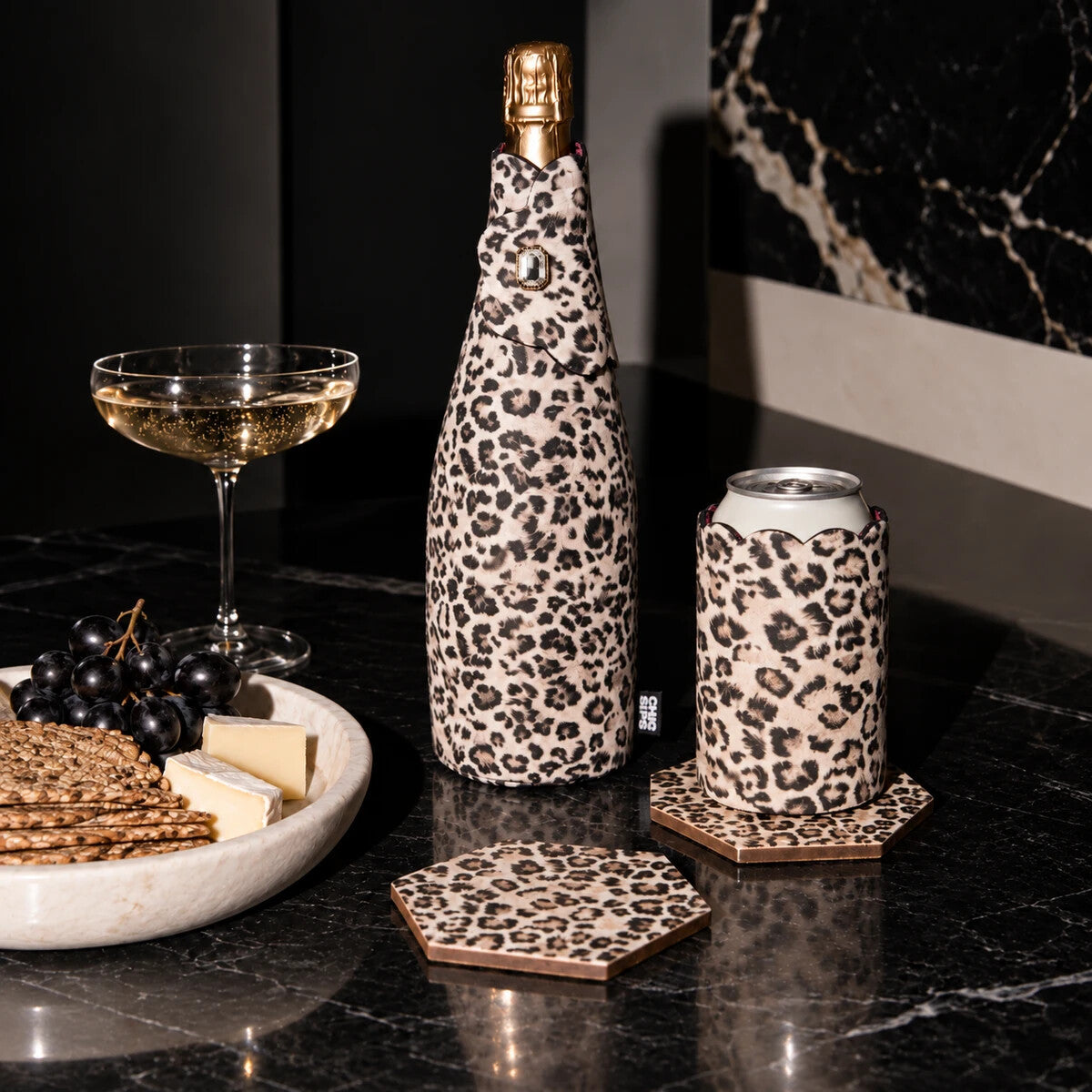 The Full Look in Catwalk leopard: a Bottle Couture jacket on a champagne bottle, a scalloped Can Couture sleeve on a can and two hexagonal Style Tiles coasters on black marble, beside a coupe of champagne and a cheese board.