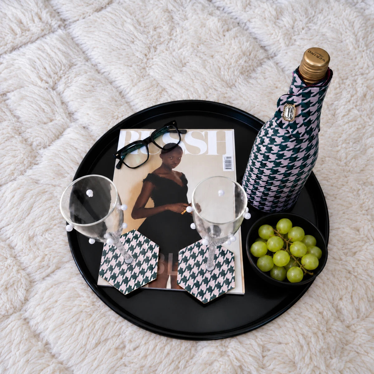 The Nightcap: a houndstooth Bottle Couture jacket on a champagne bottle and two houndstooth hexagonal Style Tiles coasters under champagne glasses on a black tray, with a magazine and green grapes on a cream shag rug.